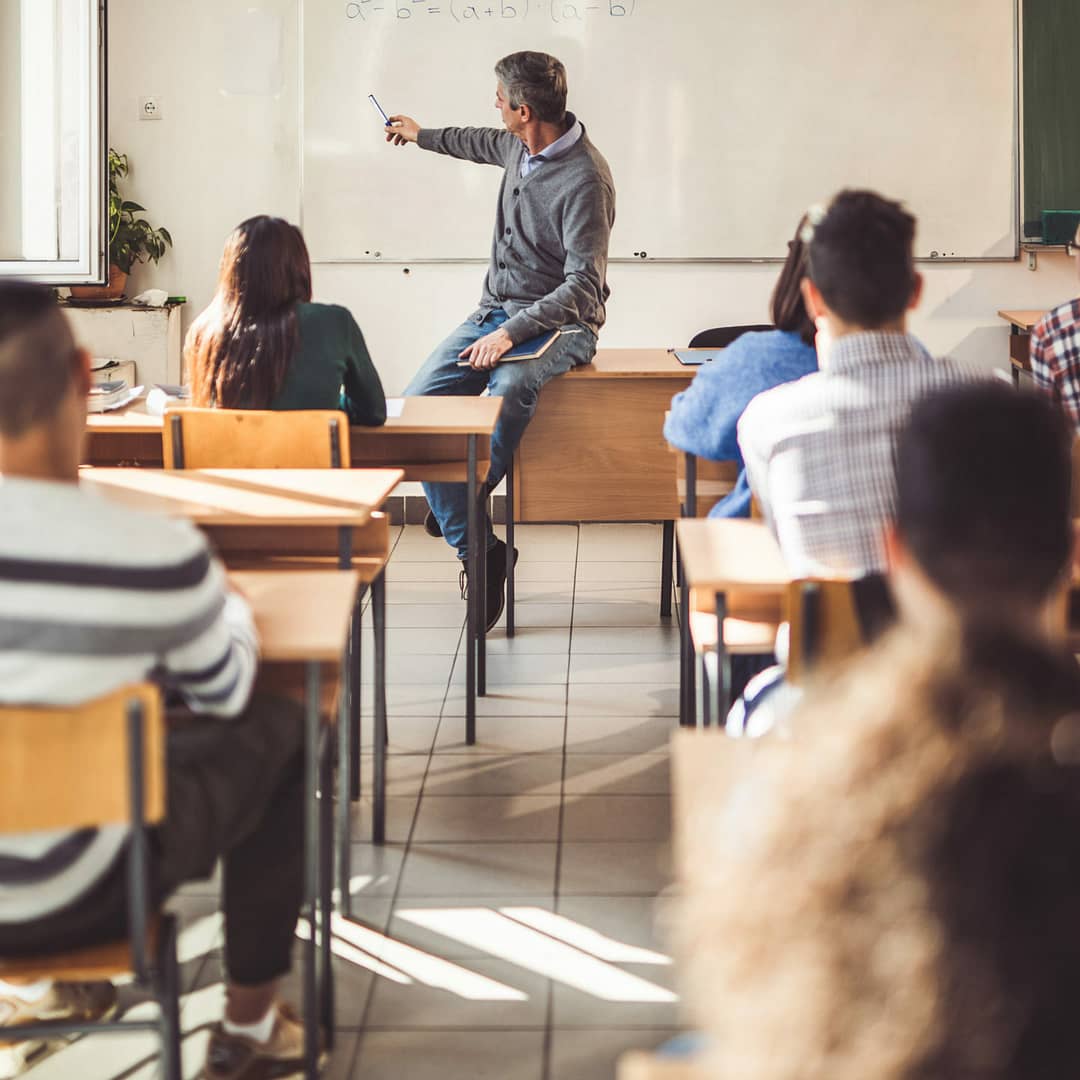 Mid adult male teacher giving a lecture to his students in the classroom. Male professor sitting on the table and teaching mathematics from whiteboard at high school.
