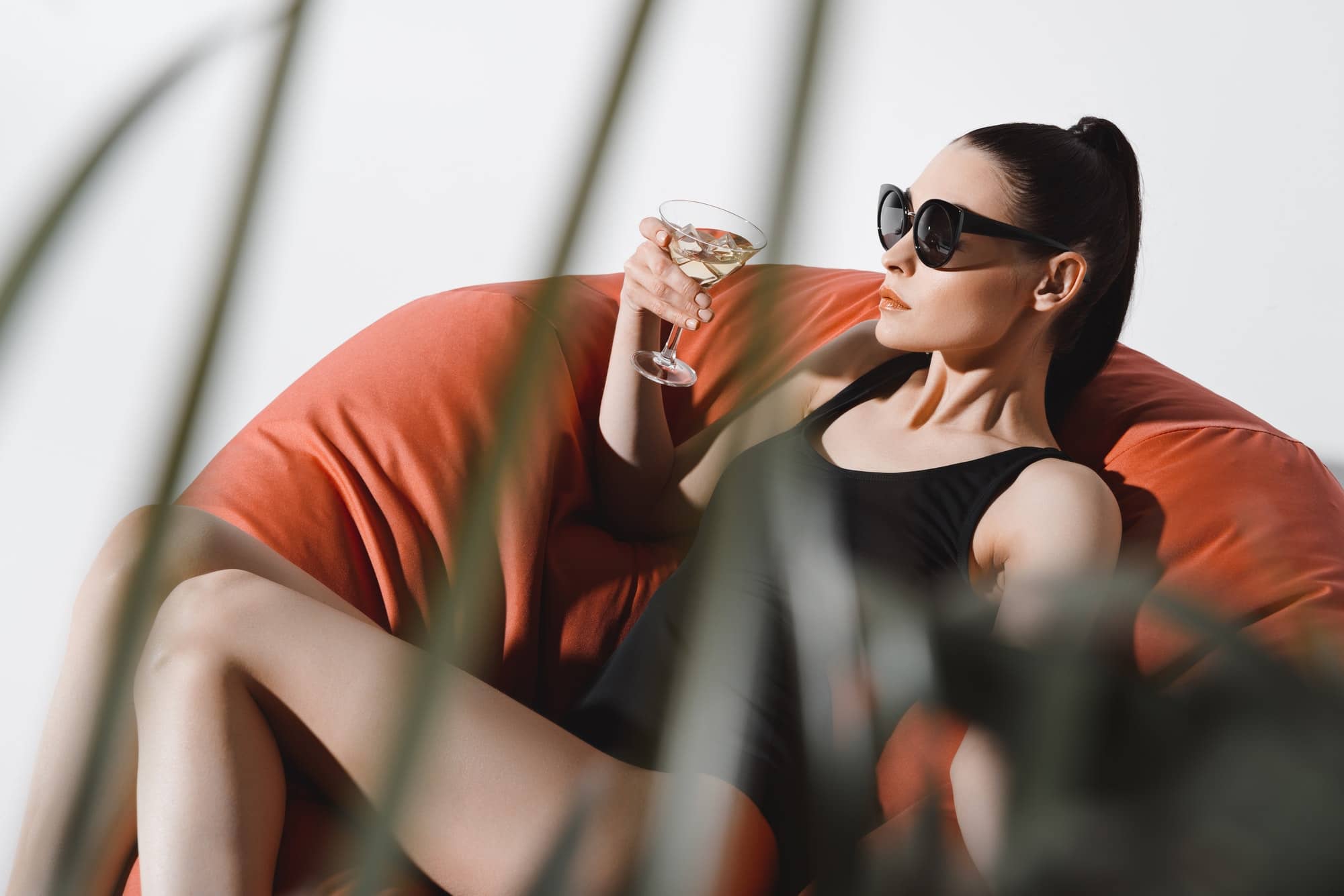 Gorgeous young woman in sunglasses and bodysuit drinking cocktail while resting in bean bag chair